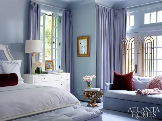 Light blue bedroom with a white dresser, a bed with white linens and a burgundy bolster pillow, and a blue chaise lounge with burgundy and pink pillows.