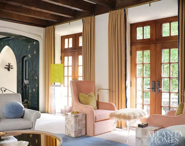 Living room with plush pink armchairs, a patterned armchair, and wooden French doors leading outside. Dark floral wallpaper adorns an arched doorway.