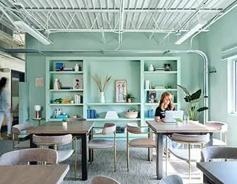 Mint green office space with built-in bookshelves, light wood tables, and upholstered chairs. A woman works on a laptop at one table.