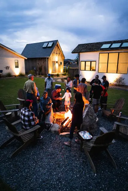 Group of people gathered around a campfire in a backyard at dusk, with modern houses visible in the background.