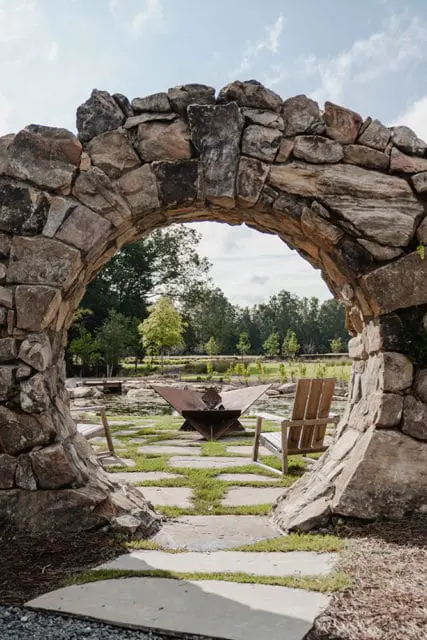 Stone archway framing a backyard with a metal fire pit, two wooden chairs, and a pond surrounded by trees.