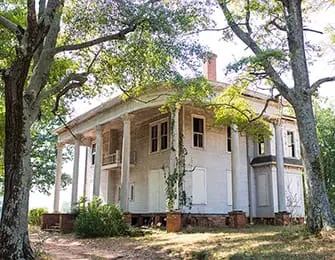 Neoclassical mansion with a wraparound porch supported by columns, surrounded by mature trees and overgrown vegetation.