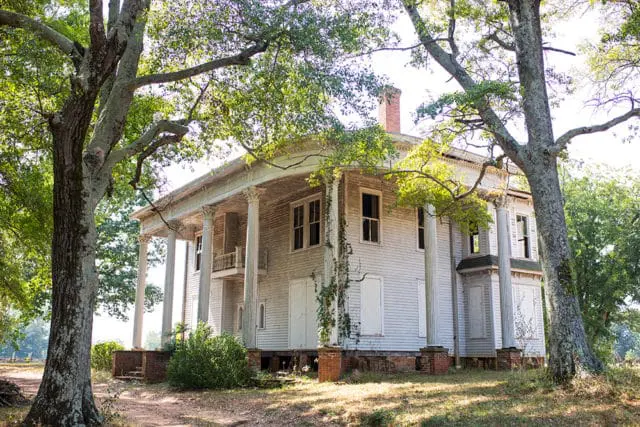 Neoclassical white wooden house with a large wrap-around porch supported by Corinthian columns, surrounded by mature trees and overgrown grass.