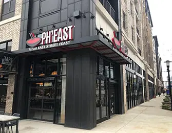 Modern building exterior with PH'EAST signage, featuring dark metal siding, glass garage doors, and a red and gray awning.