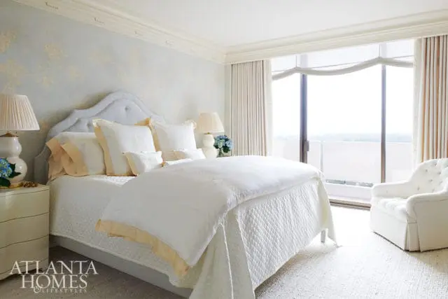 Bedroom with a tufted headboard, white bedding with pale yellow trim, and a light blue floral wallpaper. A large window overlooks an outdoor balcony.