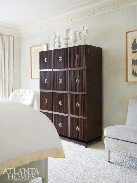 Bedroom with a dark wood dresser featuring fluted doors and brass hardware, topped with white vases and abstract art. Light blue floral wallpaper and a tufted chair are visible.
