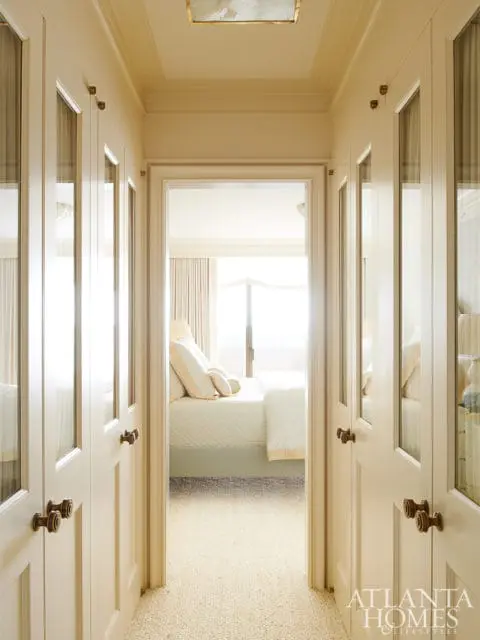 Hallway with cream-colored walls and doors featuring glass panels and brass hardware, leading to a bedroom with a bed dressed in white linens.