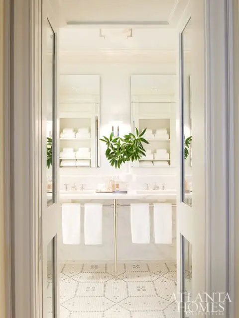 White bathroom with double vanity, marble countertops, mosaic tile flooring, and open shelving with neatly folded white towels.