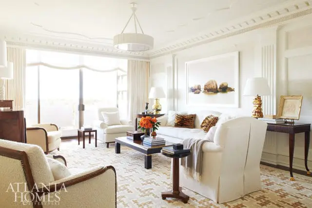Living room with white sofa, armchairs, and a patterned rug. Features a large window, artwork of rocks in water, and a dark wood coffee table.