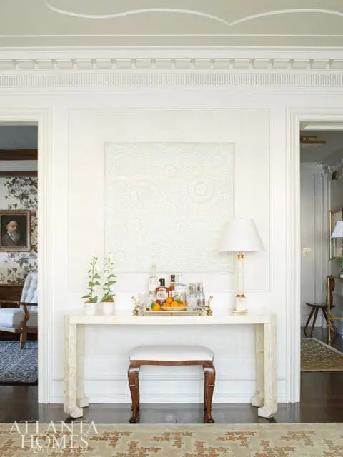 Hallway console table with marble top, bar cart, and artwork. A white upholstered stool sits in front of the table.
