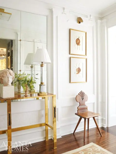 Hallway with a brass console table topped with a bust and lamp, antique wooden chair, and framed botanical prints on a textured wall.