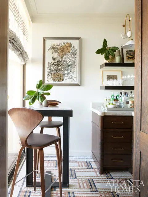 Bar area with dark wood cabinetry, marble countertop, and a high-top table with curved wooden stools. Mosaic tile flooring and framed artwork on the wall.