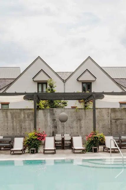 Modern white brick buildings with gabled roofs overlook a concrete wall, pool, and lounge chairs. A dark pergola with a hanging spherical light fixture is in the foreground.