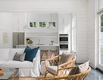 Bright living room with white shiplap walls, white sofa, rattan armchairs, and a white kitchen with stainless steel appliances and a marble island.