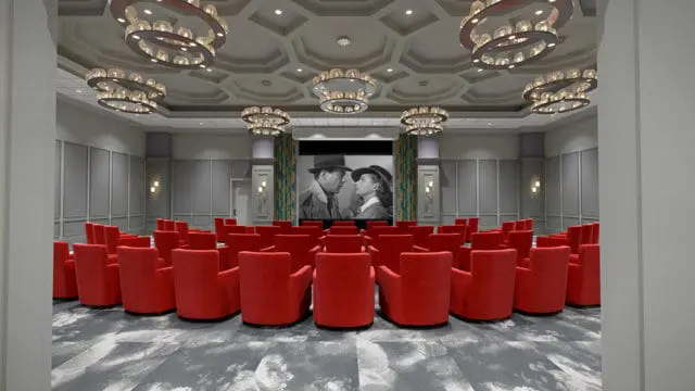 Theater room with rows of red armchairs facing a large screen displaying a black and white movie scene. Ornate circular chandeliers hang from the coffered ceiling.