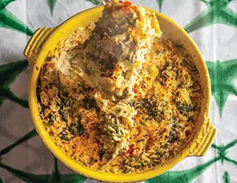 A scoop of creamy spinach artichoke dip is lifted from a yellow ceramic baking dish.