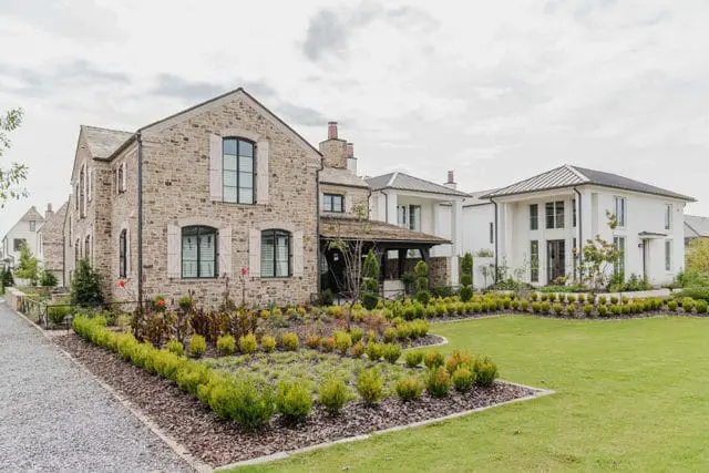 Stone-clad house with light shutters and arched windows, next to a white modern house with a metal roof, surrounded by manicured gardens and a gravel driveway.