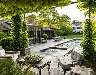 Stone patio with outdoor seating and a swimming pool and spa, surrounded by lush greenery and a pergola covered in ivy.