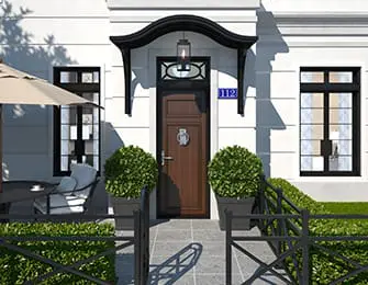 White house exterior with a dark wood front door, black awning, and two manicured bushes flanking the entrance. A metal fence and patio umbrella are visible.