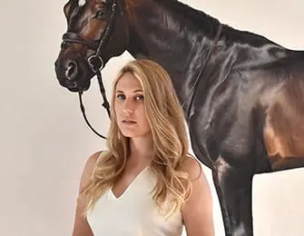 Blonde woman in a white dress stands in front of a large painting of a dark brown horse with a bridle.