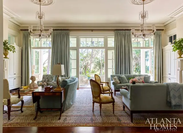 Light blue sofas and chairs arranged in a living room with tall windows and doors opening to an outdoor patio. Crystal chandeliers hang from the ceiling.