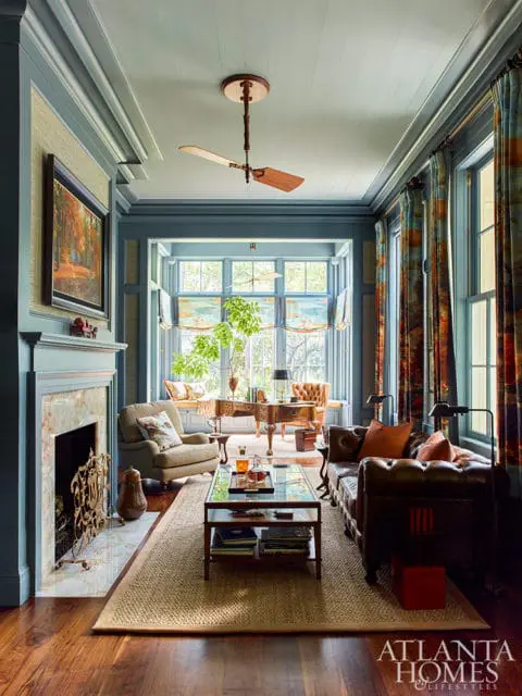 Living room with blue walls, marble fireplace, leather sofa, and patterned curtains. A wooden desk sits by a bay window.