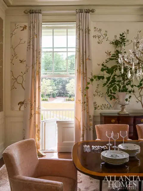 Dining room with floral wallpaper, floor-to-ceiling curtains with bird and branch motif, crystal chandelier, and a dark wood table set with plates and wine glasses.
