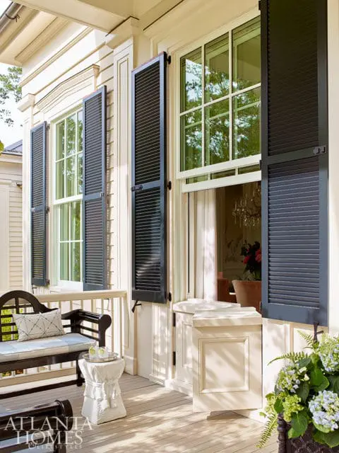 Cream-colored house exterior with black louvered shutters on multiple windows, a dark wood bench with cushions, and a white garden stool on a wooden porch.