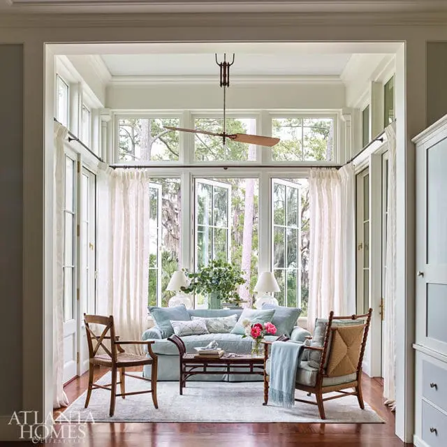 Sunroom with light blue sofa, woven armchairs, and a dark wood coffee table, surrounded by large windows overlooking trees.