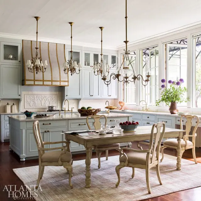 Light blue kitchen with a large island, marble countertops, brass fixtures, and a distressed wooden dining table with upholstered chairs.