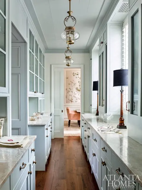 Light blue butler's pantry with marble countertops, brass hardware, glass-front cabinetry, and hanging pendant lights.