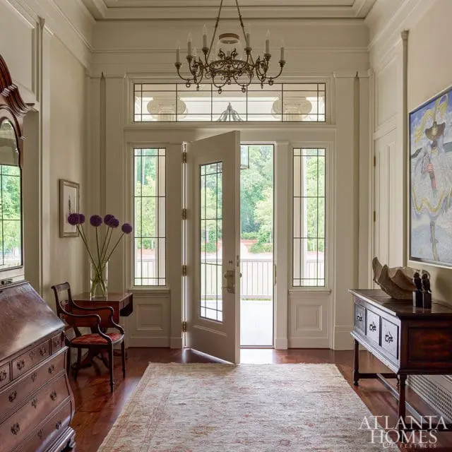 Cream-colored entryway with double doors, a chandelier, a wooden desk, a rug, and a mosaic artwork on the wall.