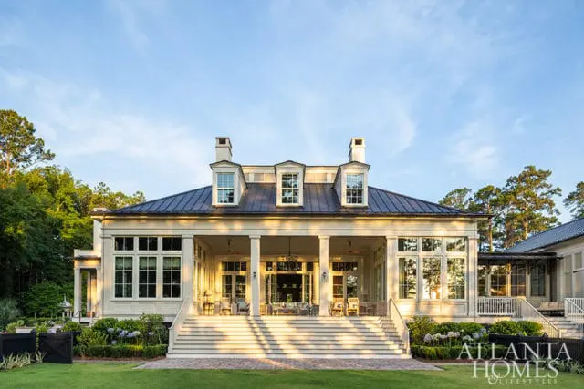 Large cream-colored house with a dark metal roof, multiple dormer windows, and a wide covered porch with seating. A grand staircase leads up to the porch, with manicured gardens and trees surrounding the property.