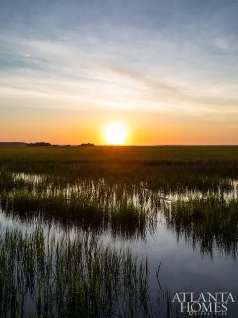 Sunset over a marsh with tall grasses reflected in the calm water. The sky is a gradient of orange, yellow, and blue with wispy clouds.