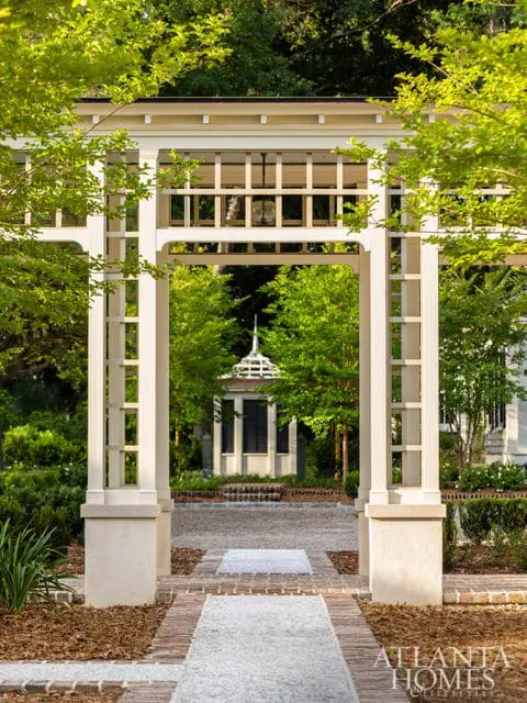 White pergola framing a view of a gazebo in a lush garden with brick pathways and mature trees