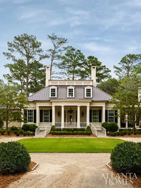 Southern-style house with a metal roof, dormer windows, white columns, and a covered porch, surrounded by manicured lawns and mature trees.