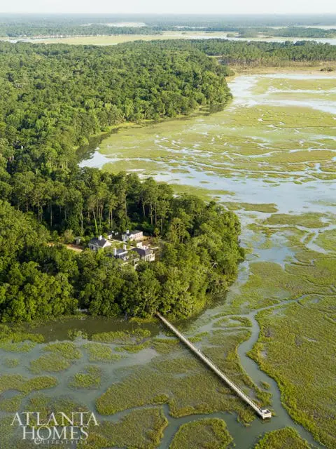 Aerial view of a white house with multiple buildings nestled in a lush green forest, overlooking a marshy waterway with a long pier extending into the water.