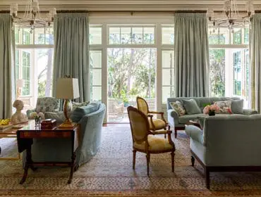 Light blue sofas and armchairs arranged in a living room with floor-to-ceiling windows overlooking a garden. Features a patterned rug, crystal chandeliers, and built-in cabinetry.