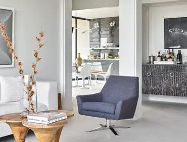 Living room with white sofa, blue armchair, and a bar cabinet with bottles and glassware. A doorway reveals a kitchen with white tulip chairs.