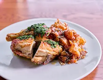 Roasted chicken quarter topped with green herb sauce served with crispy fried potatoes on a white plate