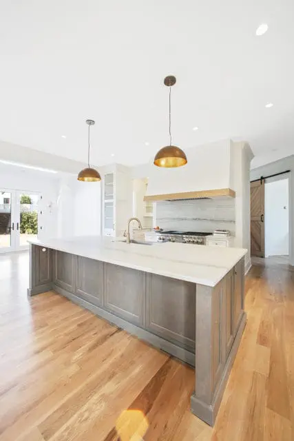 Gray kitchen island with white marble countertop and brass pendant lights, overlooking a stainless steel range with marble backsplash and a wooden barn door.