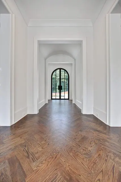 Hallway with white walls and arched doorway featuring a black metal door, leading to a view of trees. Dark wood herringbone floors run the length of the hallway.
