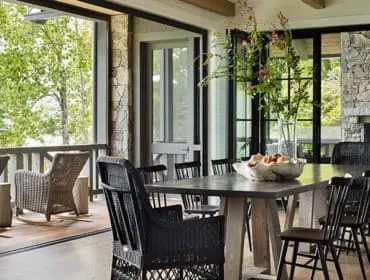 Dining room with a large stone fireplace, dark wood dining table set with black chairs, and wicker rocking chairs on a screened porch overlooking a lake.
