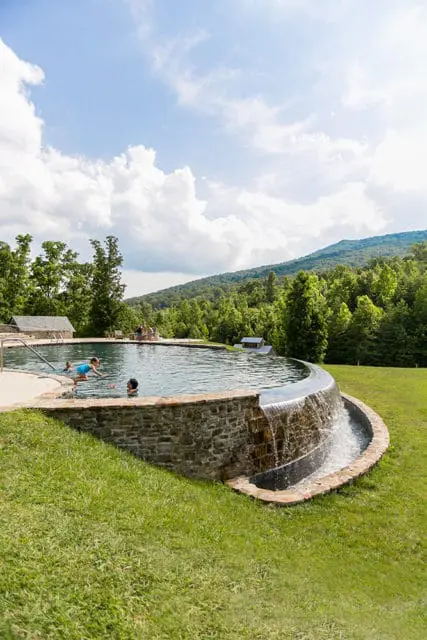 Curving infinity pool with stone retaining wall and waterfall feature, surrounded by green grass and lush trees with mountains in the background under a cloudy sky.