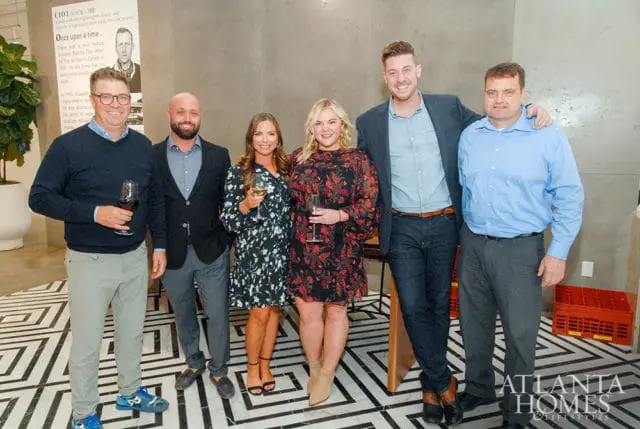 Six people stand in front of a wall with text and a portrait. The floor has a black and white geometric pattern. The Atlanta Homes & Lifestyles logo is visible in the lower right corner.