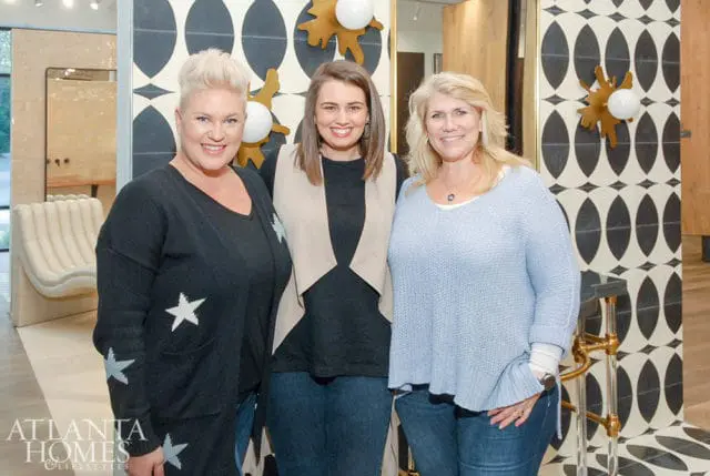 Three women smiling at the camera in front of a patterned wall. The woman on the left wears a black cardigan with white stars. The woman in the middle wears a black shirt and a tan vest. The woman on the right wears a light blue sweater.