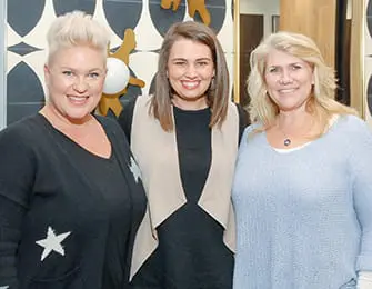 Three women smile at the camera. The woman on the left wears a black sweater with white stars. The woman in the middle wears a tan vest over a black top. The woman on the right wears a light blue sweater.