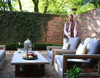 Woman standing on a gravel patio with brick walls, ivy-covered wall, and outdoor seating area with a coffee table and chairs.