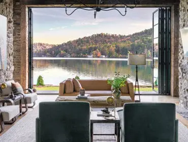 Living room with stone walls, featuring a tan leather sofa, two green armchairs, and a large fireplace. Bi-fold doors open to a lake view with tree-covered hills.