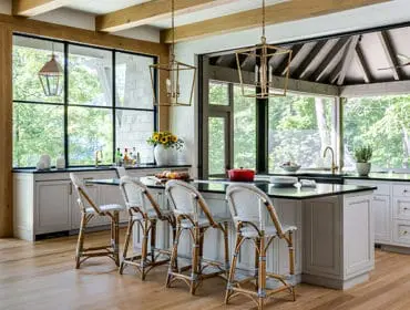 Kitchen with light wood floors, white cabinetry, black countertops, and a large island with rattan bar stools. Large windows overlook a wooded area.